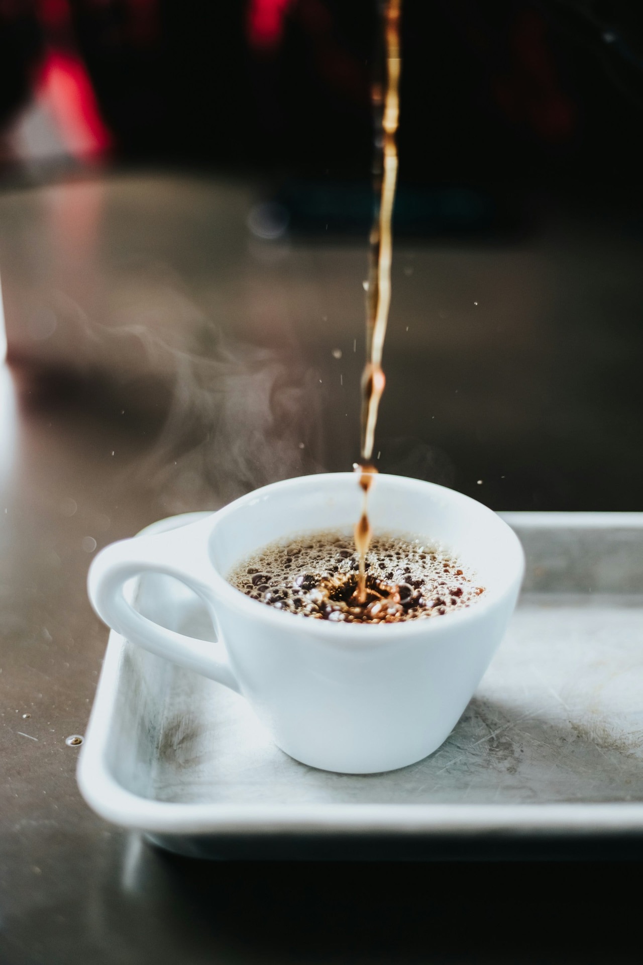 Wit kopje met oortje op een metalen serveerplaatje. Het kopje wordt gevuld met koffie, er valt een straal in.