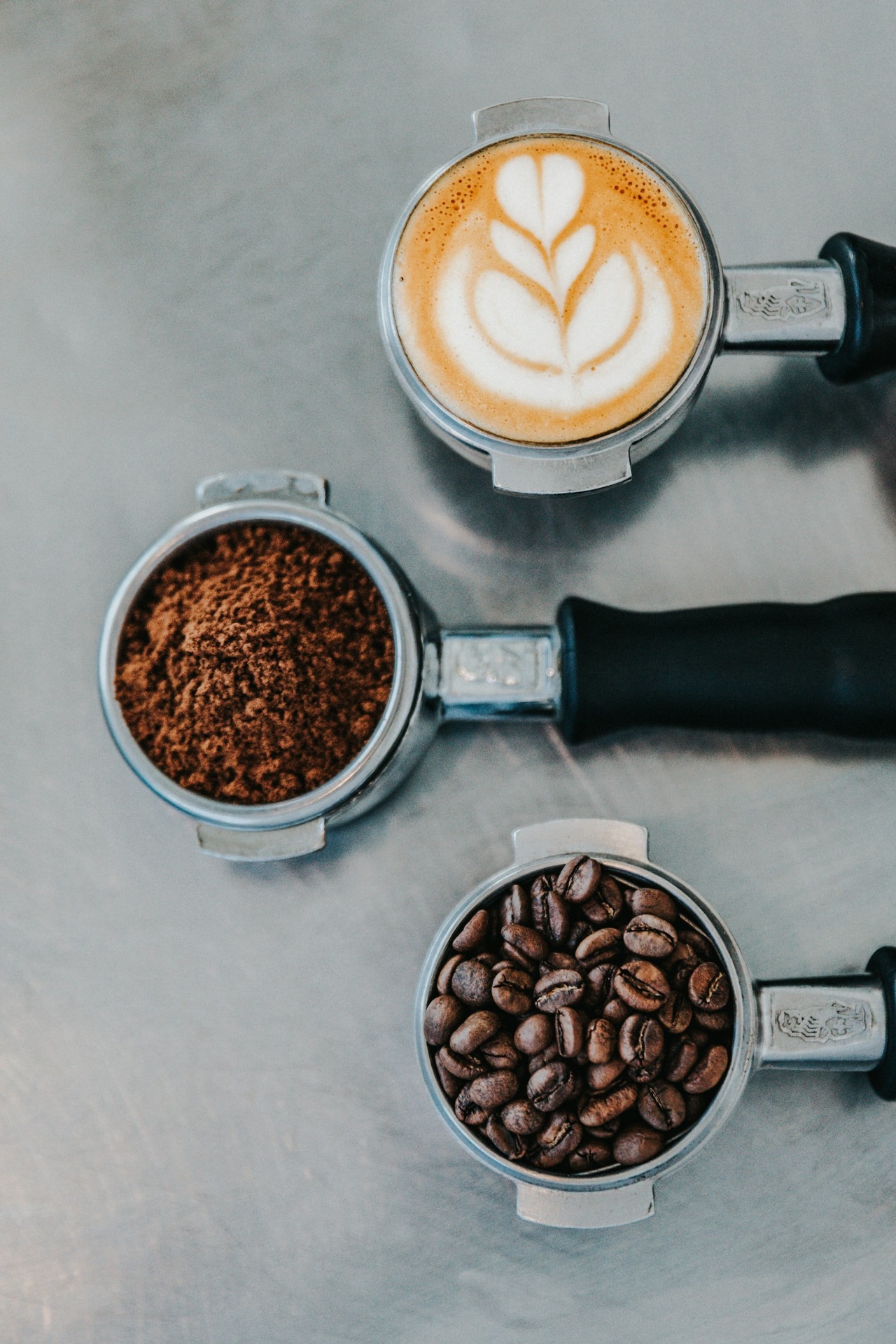 Drie koffiepistons op een metalen ondergrond. De middelste steekt iets verder uit dan de buitenste twee. In de linker piston zitten bonen, in de middelste gemalen koffie en in de rechter piston zit een cappucinovulling.