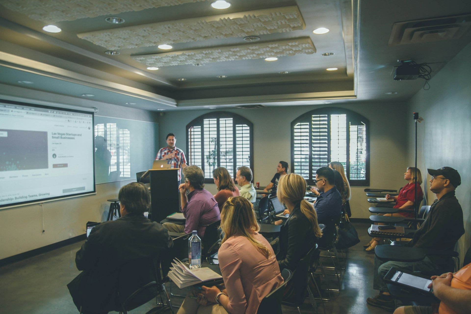 Een ruimte met meerdere volwassen mensen die luisteren naar een man die voorin de ruimte staat en naast een projectiescherm iets uitlegt. Op het scherm staat de tekst: Las Vegas Startups and Small Businesses.