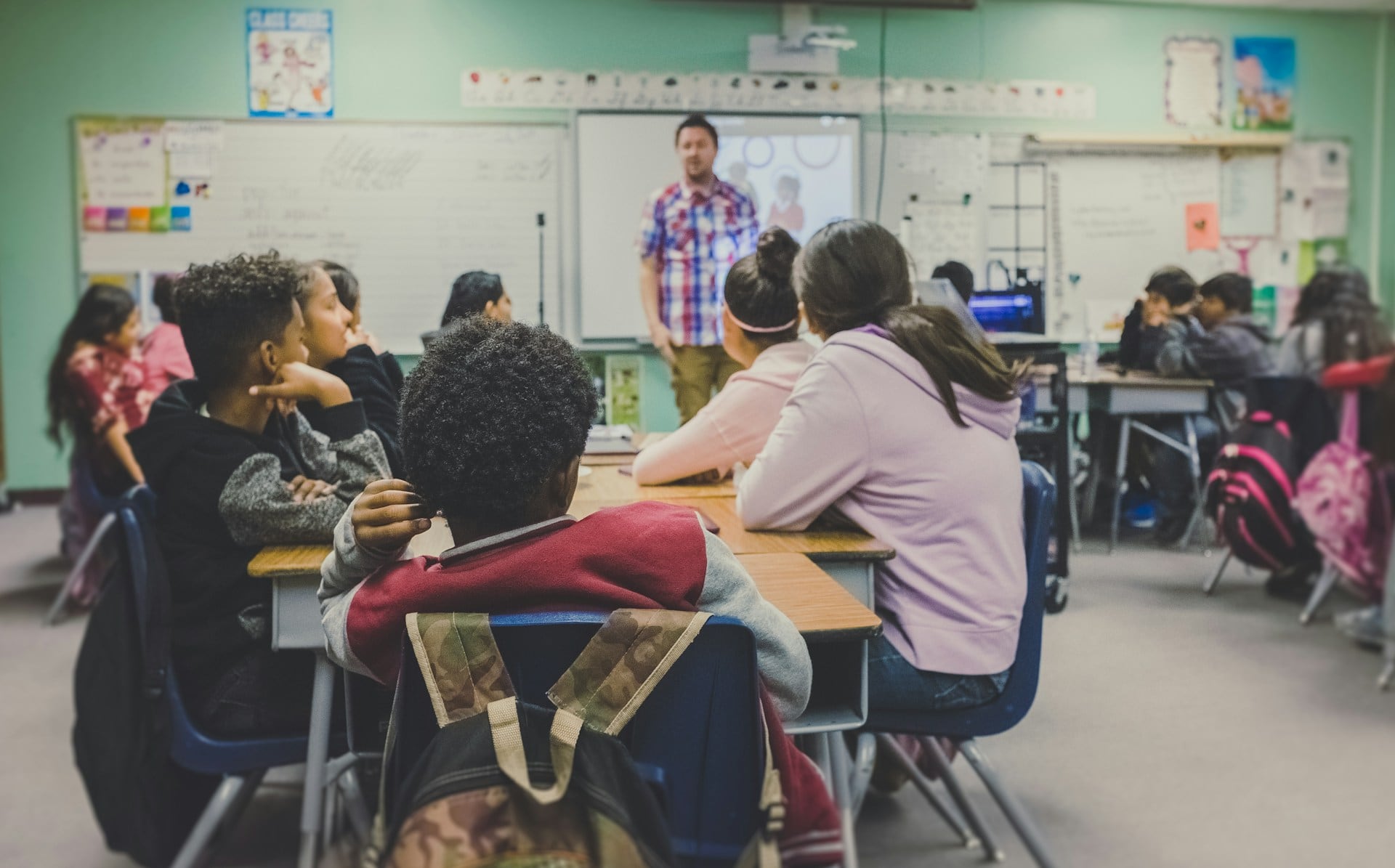 Een klaslokaal met kinderen die luisteren naar een docent, een man die voor een whiteboard staat. De man heeft een geruit overhemd met korte mouwen aan.
