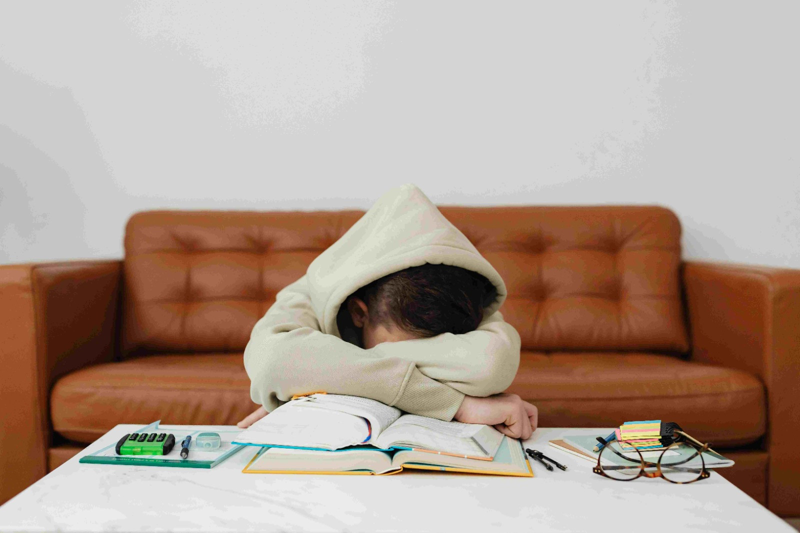 Bruin leren bank met op de voorgrond een witte tafel met een kind in een beige hoodie, heeft de capuchon half op, kort zwart haar steekt eronderuit. Heeft zijn armen over elkaar en zijn gezicht omlaag. Zijn armen rusten op twee opengeslagen boeken. Naast hem ligt een passer, een rekenmachine en een kladblok en op de voorgrond een bril