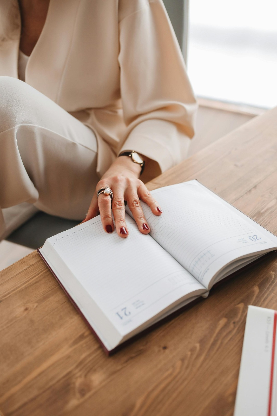 Een dameshand met roodgelakte nagels rust op een opengeslagen agenda. Om de wijsvinger worden twee ringen gedragen en om de pols een horloge. De persoon draagt een beige-kleurige blouse en broek.