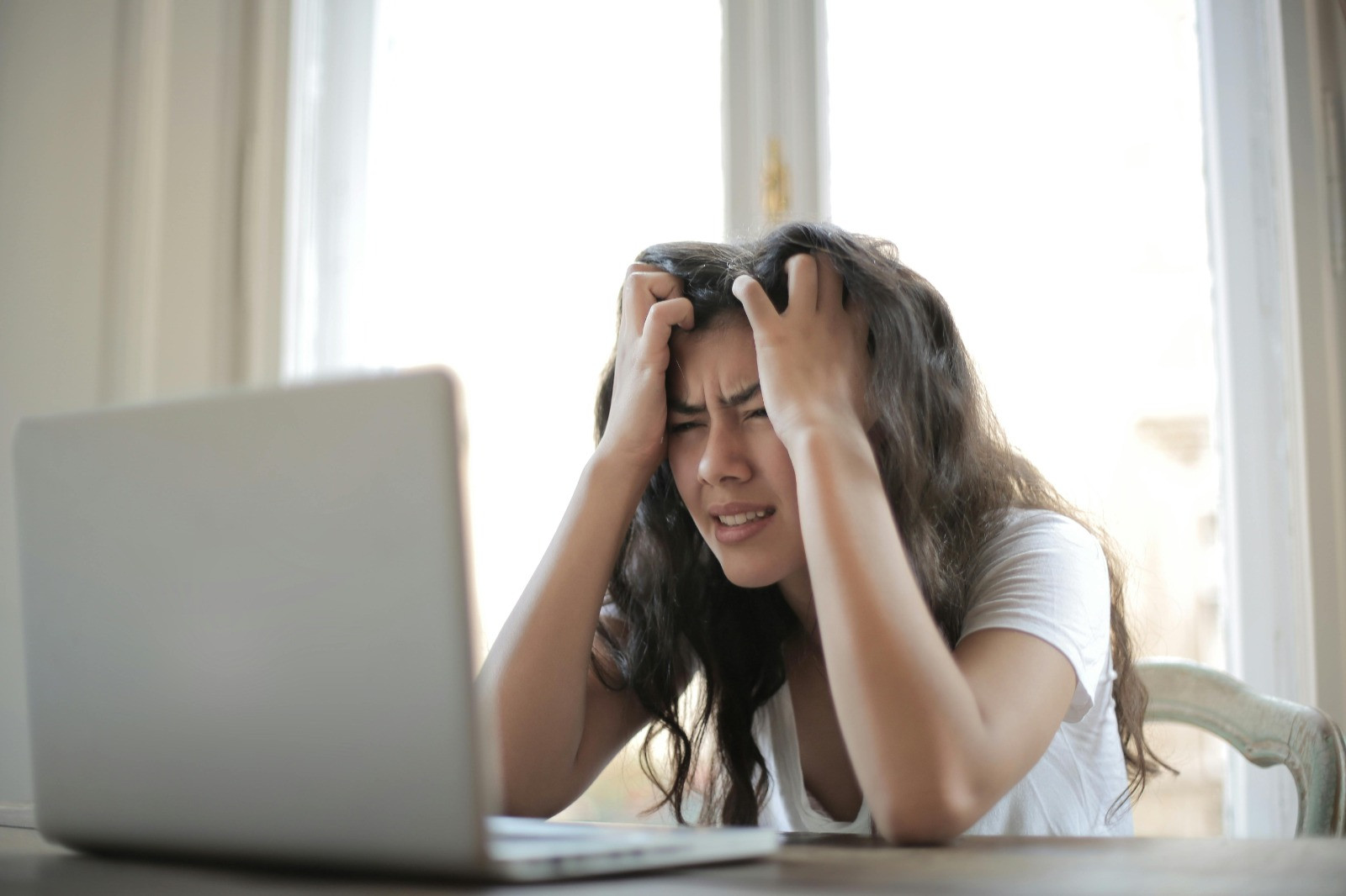 Meisje met lichtbruin haar en wit T-shirt zit met haar handen in het haar achter een laptop. Ze kijkt gestrest.