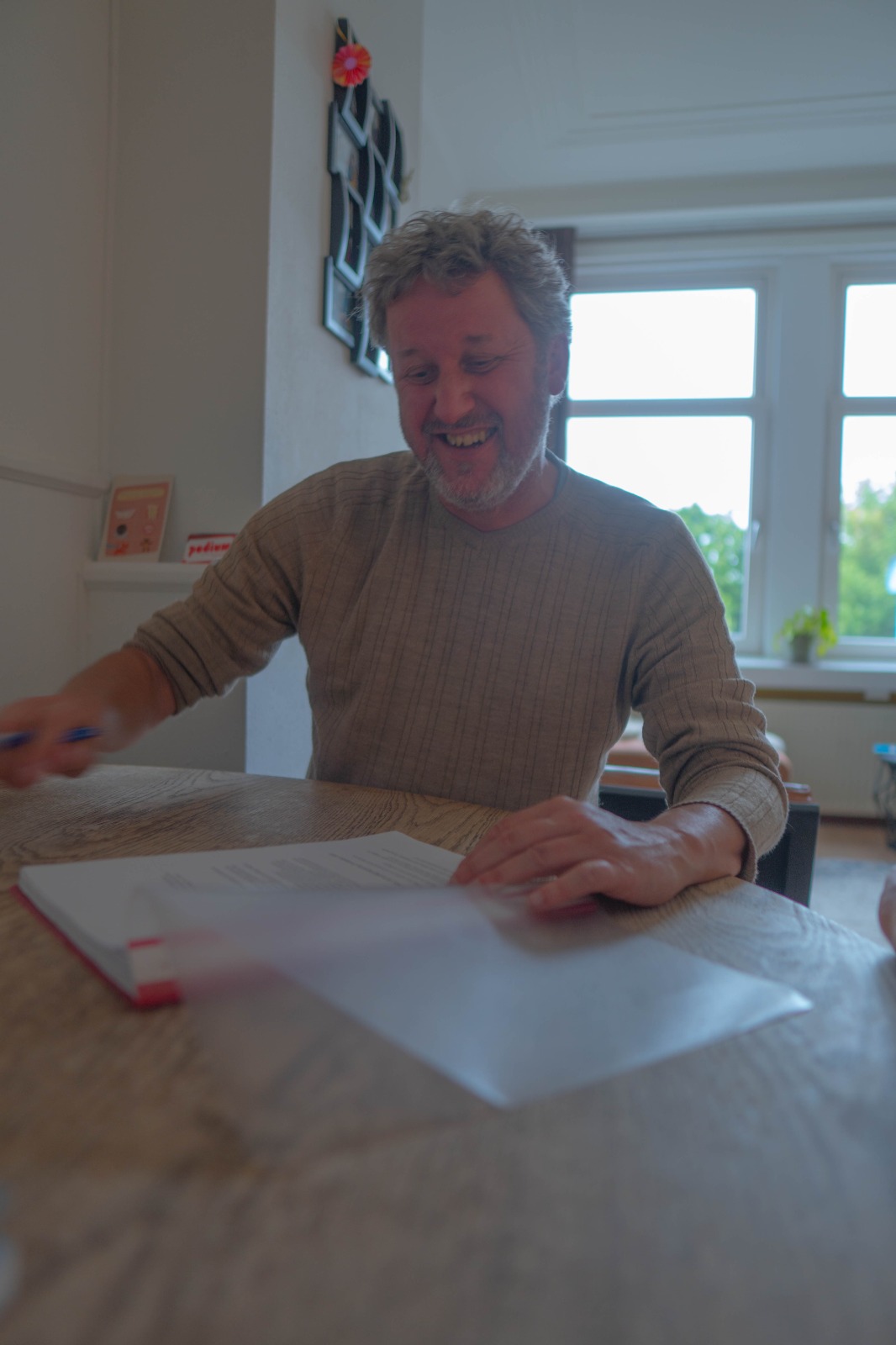 Man met grijs haar en een baard in een lichtbruine trui zit aan een houten tafel met voor zich een mapje. In zijn rechterhand heeft hij een pen vast, en achter zich zijn twee raamkozijnen te zien.