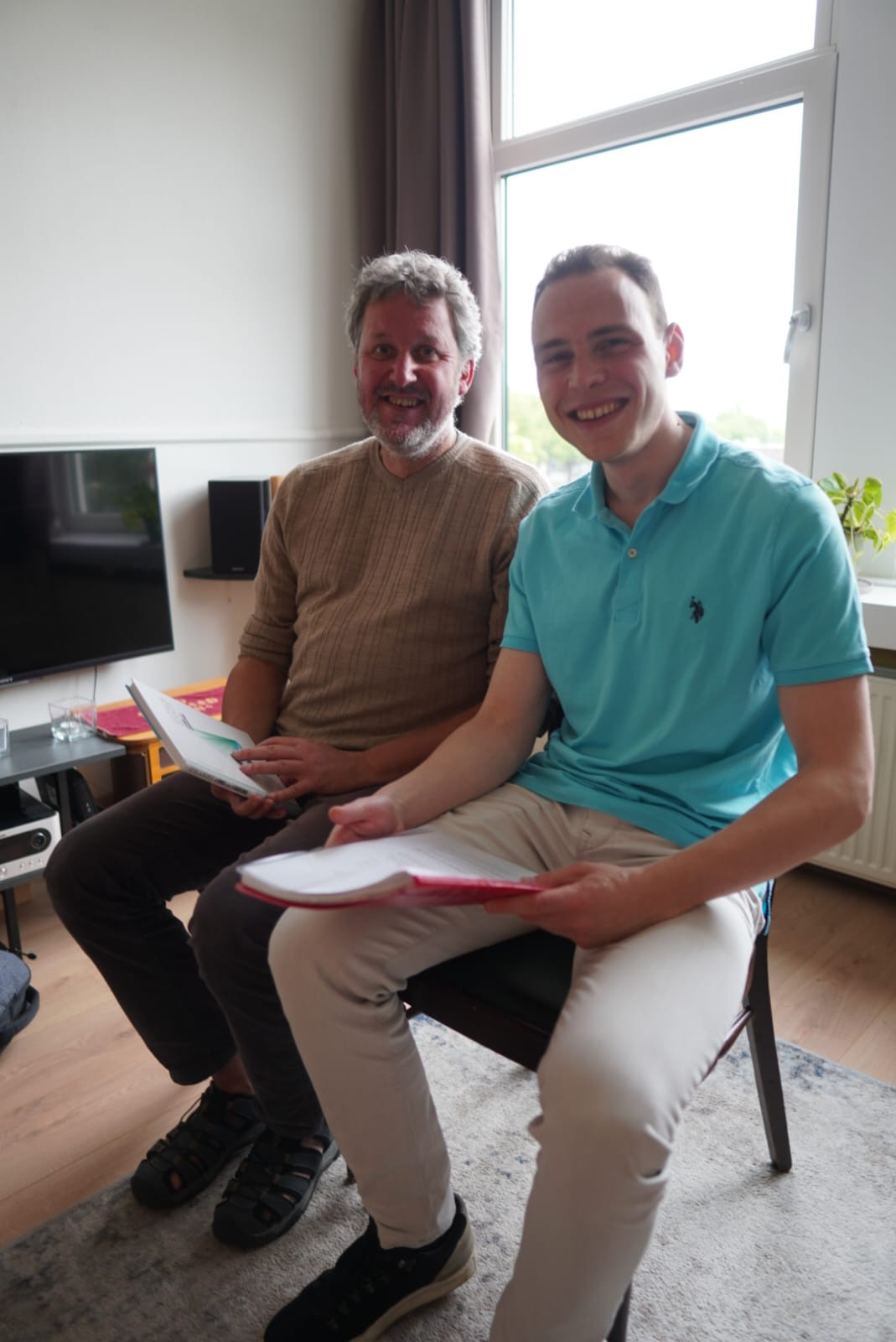 Twee personen zitten naast elkaar op een stoel, een jongere en oudere man. De jongere persoon heeft kort, donkerbruin haar en een lichtblauwe polo aan en houdt een mapje in zijn hand, de oudere man een lichtbruine trui en een boek.