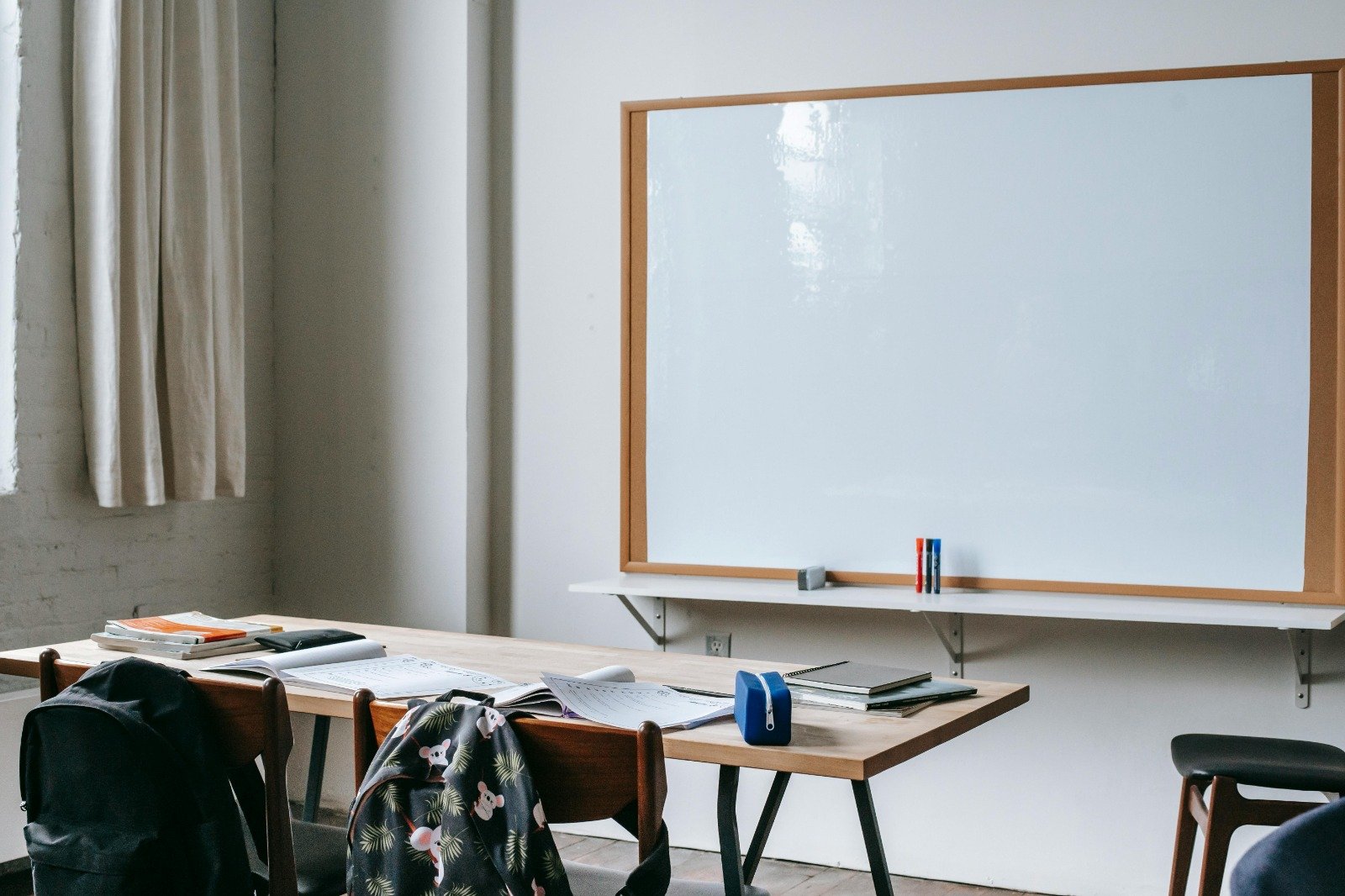 Een ruimte lijkend op een klaslokaal met vooraan een whiteboard met houten frame erom. Ervoor staan drie stiften rechtop in de kleuren rood, zwart en blauw (van links naar rechts). Op de voorgrond is een houten tafel te zien met twee stoelen eraan, waaraan rugtassen hangen. Op de tafel liggen schriften, boeken en etuis. Er lijkt te worden gewerkt door leerlingen.