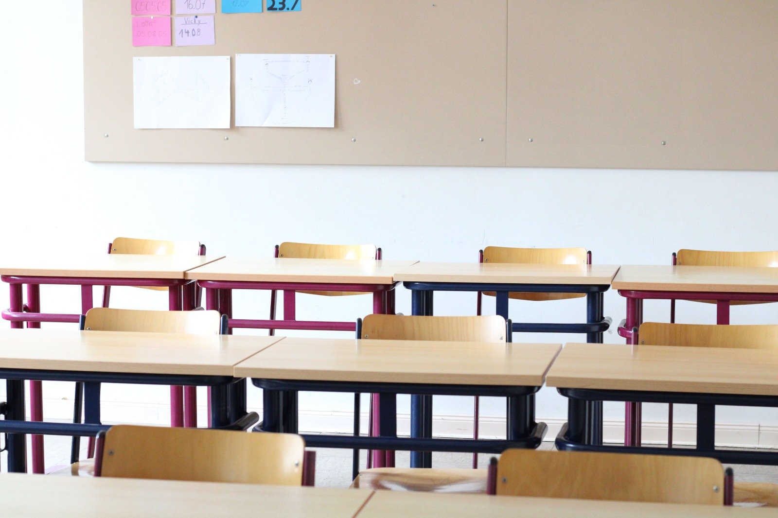 Schooltafels met stoelen in een leeg lokaal. Op de achtergrond een witte muur en een grijs prikbord.